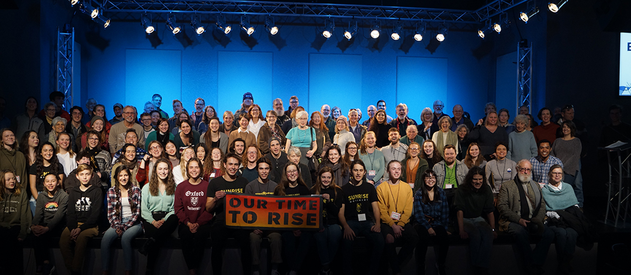 150 people gathered for a group photo with a banner saying "Our time to rise."
