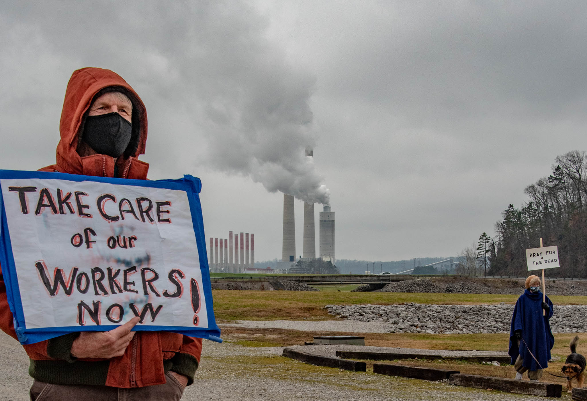 Person holding sign saying "Take Care of Our Workers Now" in front of power plant