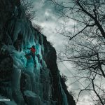 a man in a red jacket climbs a blue-tinged column of ice under a cloudy sky