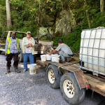 Two men stand on a gravel road outdoors next to a truck and another man crouched next to white buckets.