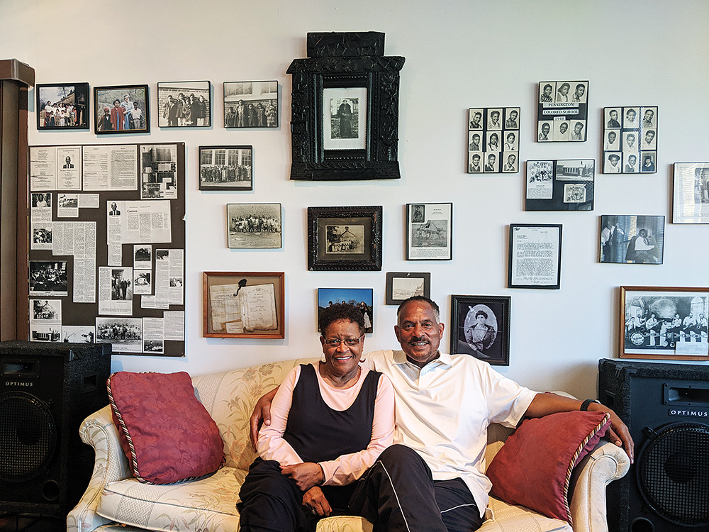Two people sit on couch in front of wall of photos and documents