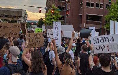 Crowd of people in a city intersection with signs saying "Change," "Stand Up, and "Know Justice, Know Peace"