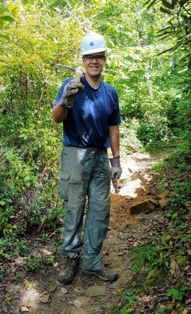 Wearing a hard hat, Dan stands in the middle of a trail with greenery on either side, holding a trail-building tool over his shoulder