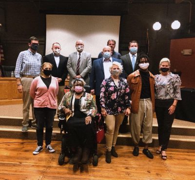 11 people, wearing masks, in the center of a meeting room