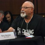 Black man with gray beard wearing a "Black Lung Kills" t-shirt speaks into a microphone, tears on his face