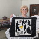 woman with light blonde hair and glasses holds a quilt square with cutout images of miners, a canary, mining boots, coal and a Bible