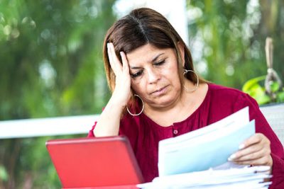 A mature hispanic woman is sitting at the dining table using her tablet to sort her household bills online.She is looking glum as she struggles to sort the family finances. She is holding an invoice. The tablet is surrounded by various bills and debt letters.