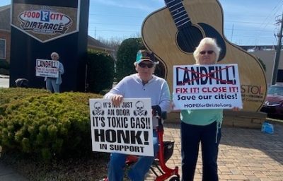 two people in front of a large guitar statue hold signs about the landfill problems