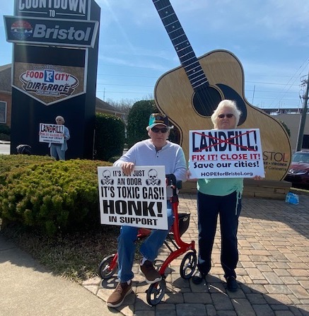 two people in front of a large guitar statue hold signs about the landfill problems 