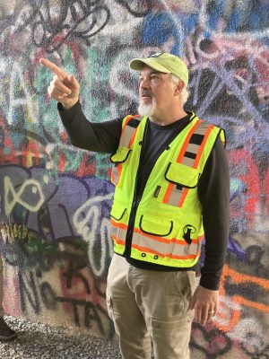 A man in a neon yellow vest stands in front of a graffiti wall pointing in the distance. 