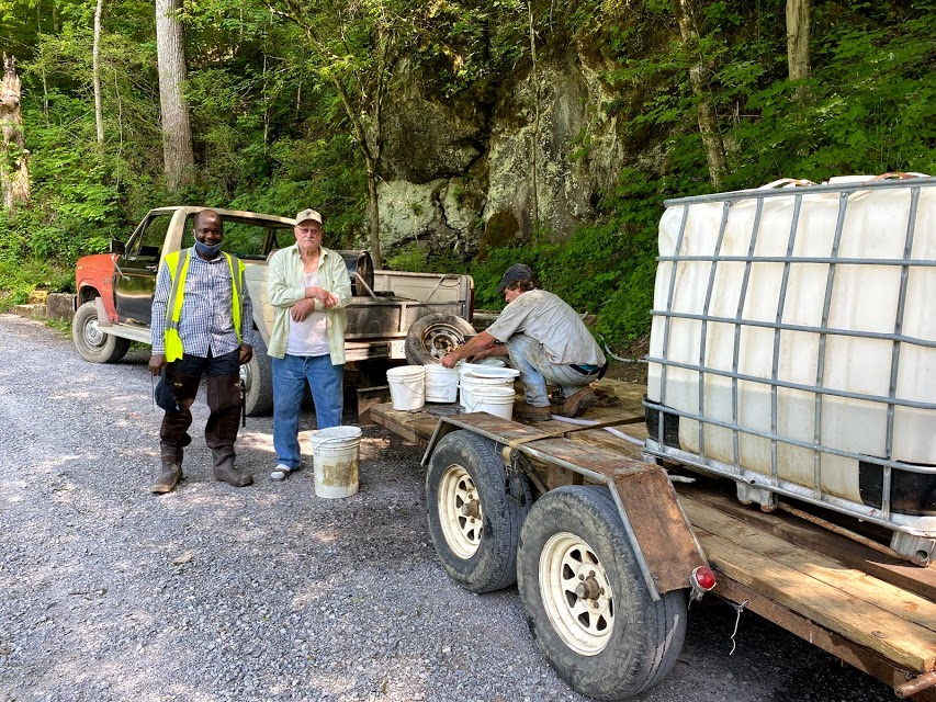 Two men stand on a gravel road outdoors next to a truck and another man crouched next to white buckets.