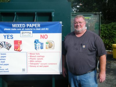man with gray hair, wearing a t-shirt stands next to a sign that describes types of mixed paper that can be recycled