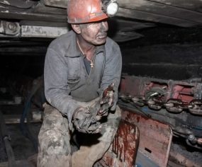 A coal miner, covered in coal dust, stoops inside a mine shaft to repair a combine.