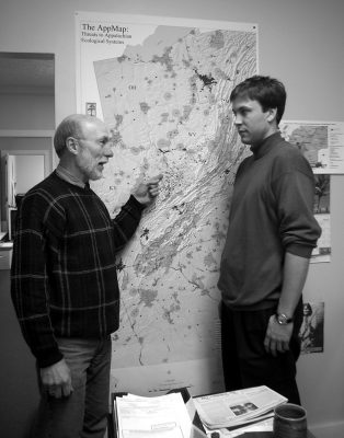 Older man on the left points at a spot on a large map, talking to a younger man who is also in front of the map