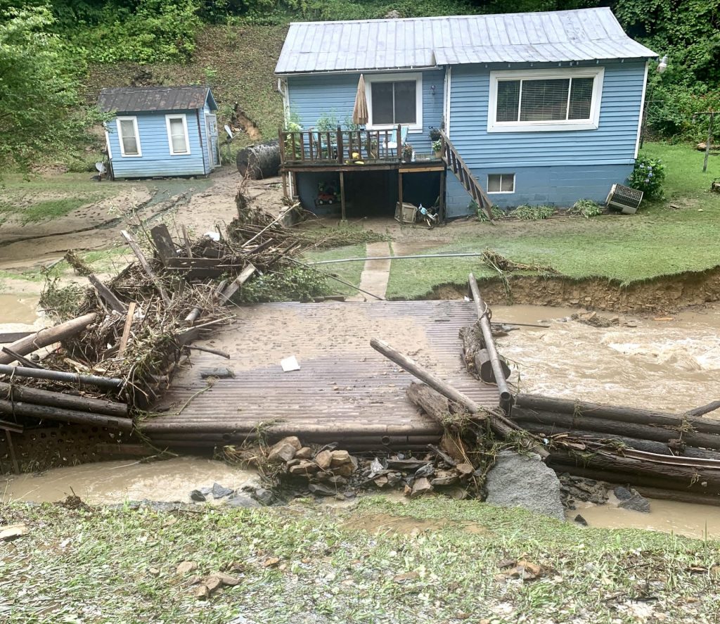 Debris covers one side of a damaged bridge that once connected a blue house to the roadway in the foreground. Receding water reveals badly eroded creek banks.