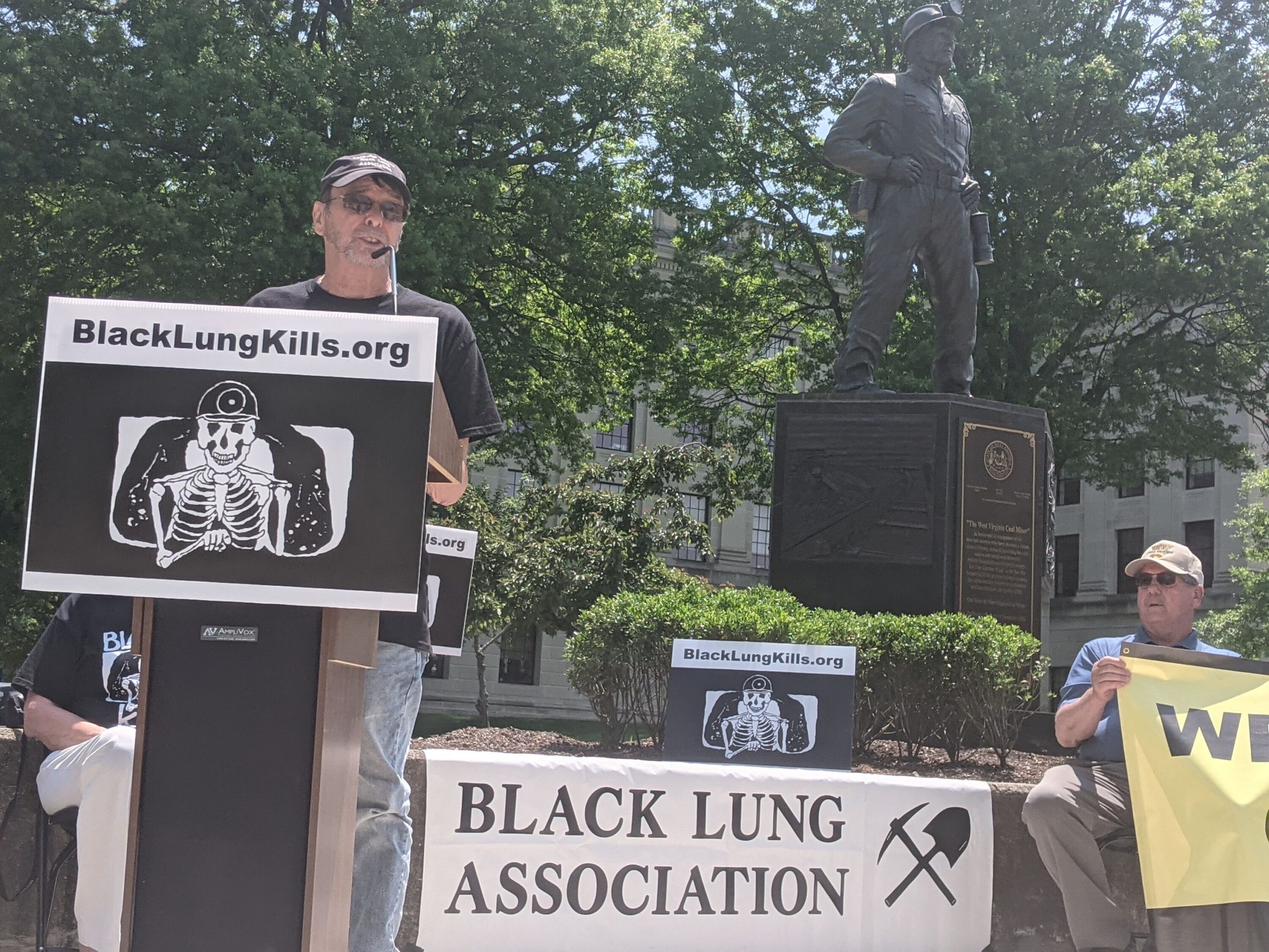 man stands at podium in front of statue of a miner, banner in background says "Black Lung Association"