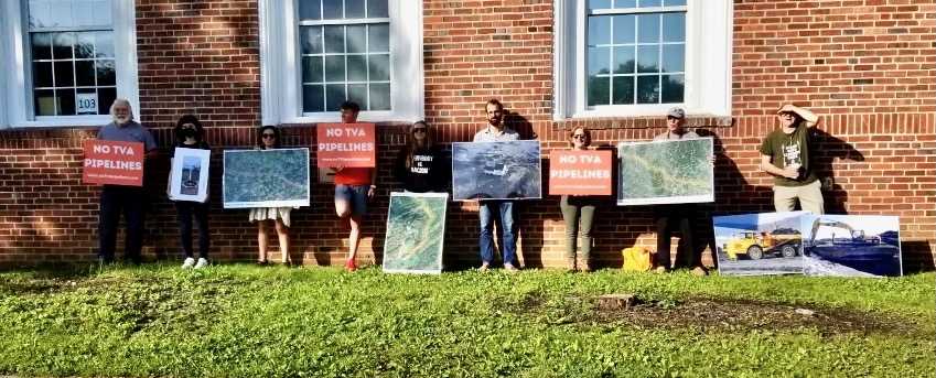 Six people stand on a lawn outside a brick school building, holding signs to protest pipelines in Tennesssee.