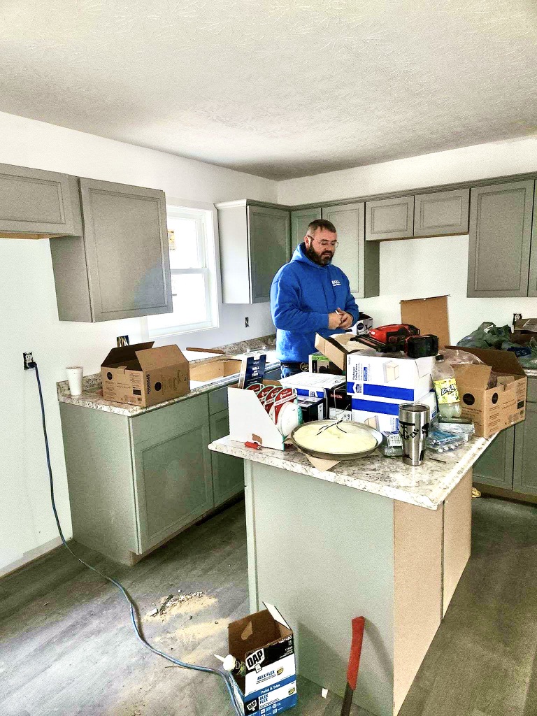 A man wearing a bright blue sweatshirt works in a kitchen amid building supplies and light fixtures, putting the finishing touches on the recently built house.