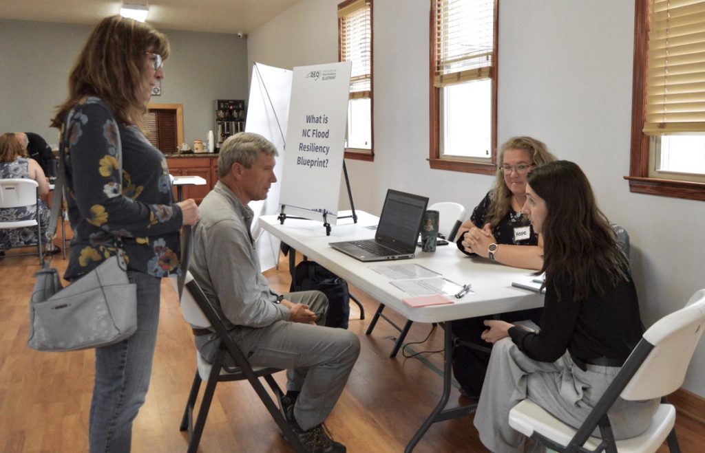A woman stands behind a man seated in a chair, while the two speak with two women seated at a long table. A laptop computer with information about flood resiliency is open on the table.