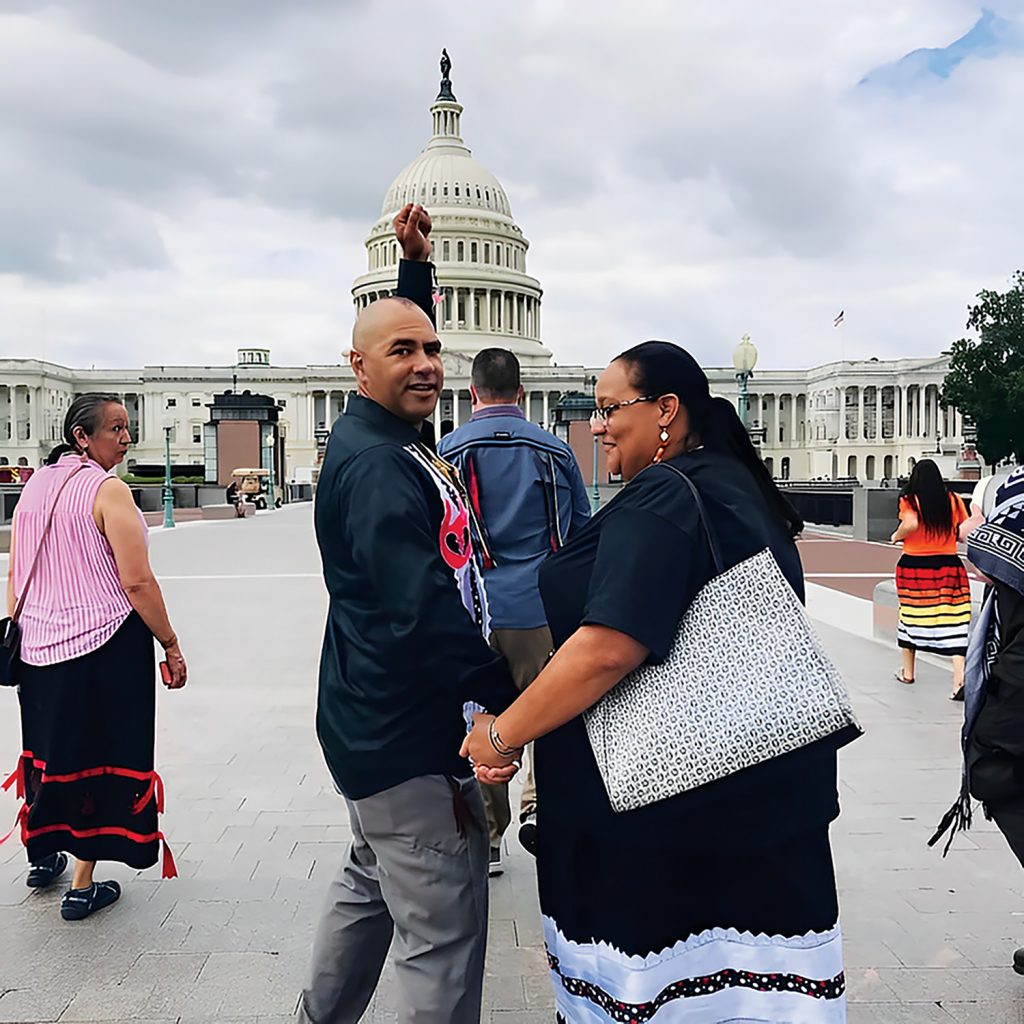An Indigenous couple stands with the Capitol Building in the background. They are smiling and the man has his fist raised above his head.