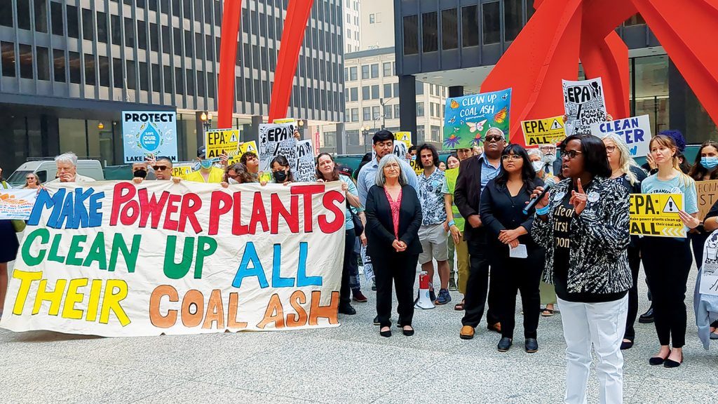A black woman speaks into a microphone. People stand behind her holding signs calling for coal ash cleanup. A large banner held by several people at the front of the group reads, "Make Power Plants Clean Up All Their Coal Ash."