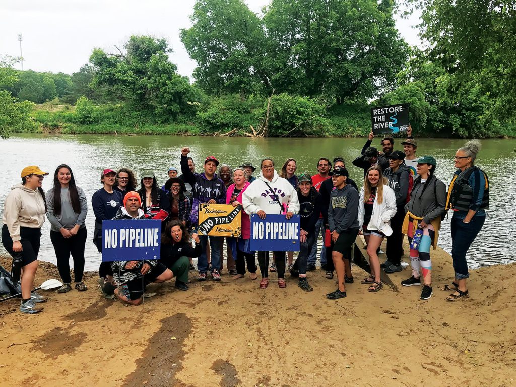 A group of people stand on a river bank, some holding signs that say "NO PIPELINE" and "RESTORE THE RIVER."