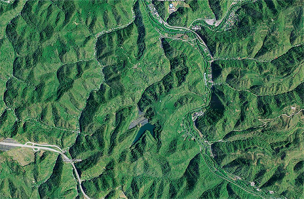 An aerial satellite image shows roads and homes below a large pond and gray earthen dam in a mountainous area.
