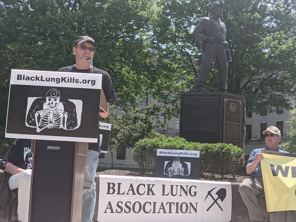 A man in a black t-shirt speaks at a podium behind a sign depicting a skeleton wearing a miner's hat that says BlackLungKills.org in black lettering. A banner that says Black Lung Association is draped over a table behind him and a statue of a miner rises above them in the background.