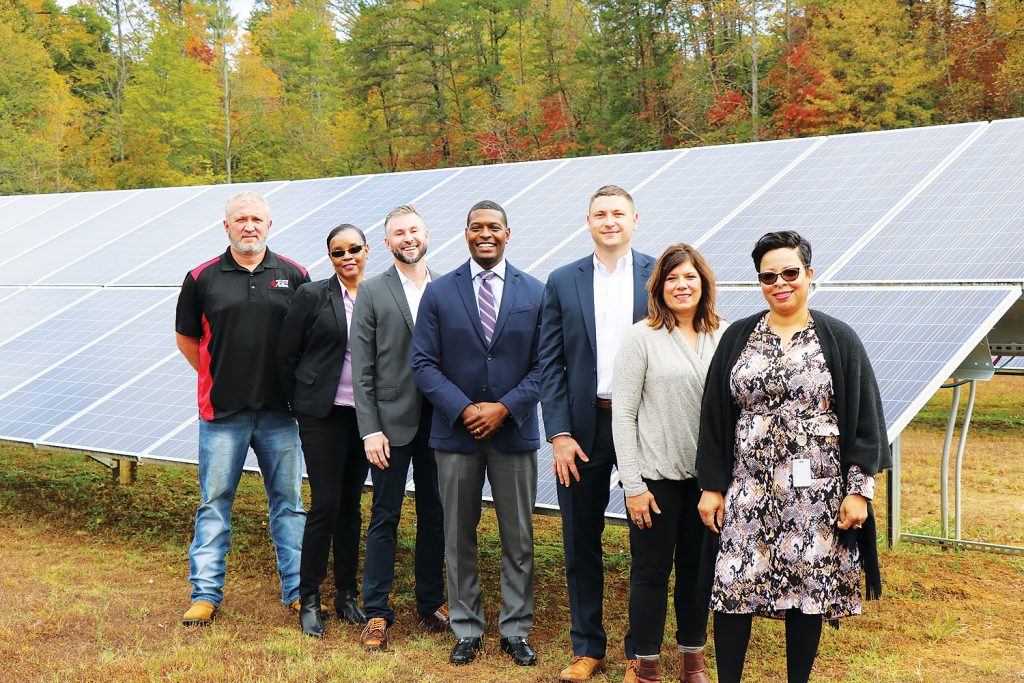 Seven people stand in front of a large solar panel array in a grassy area with trees behind it.