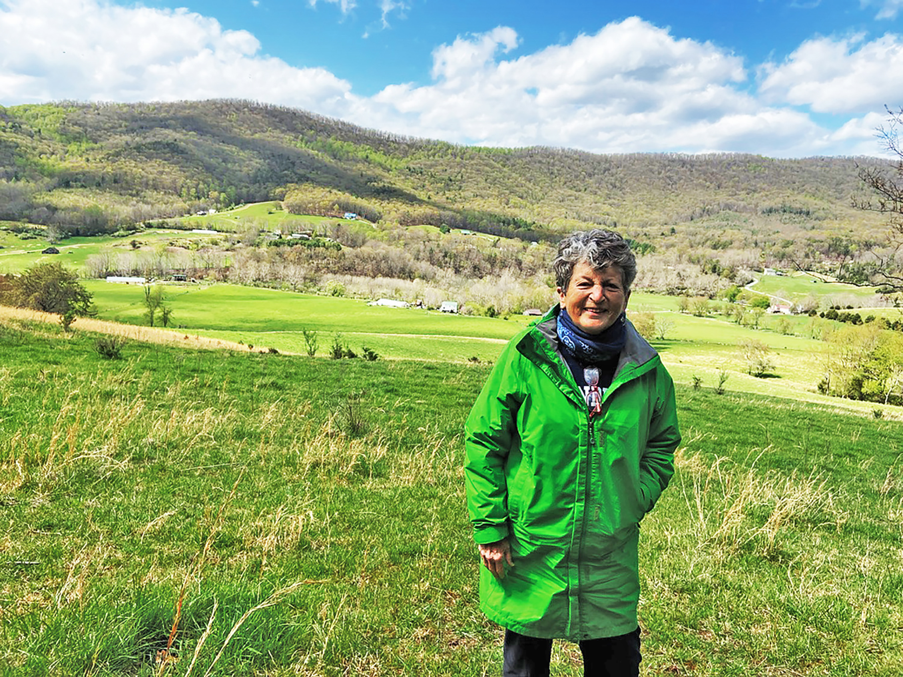A woman with short, graying hair wearing pants and a long, green jacket stands in a pasture with fields and rolling hills behind her.
