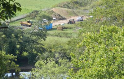 Heavy equipment and vehicles are seen in a fenced off area of disturbed land next to a river.