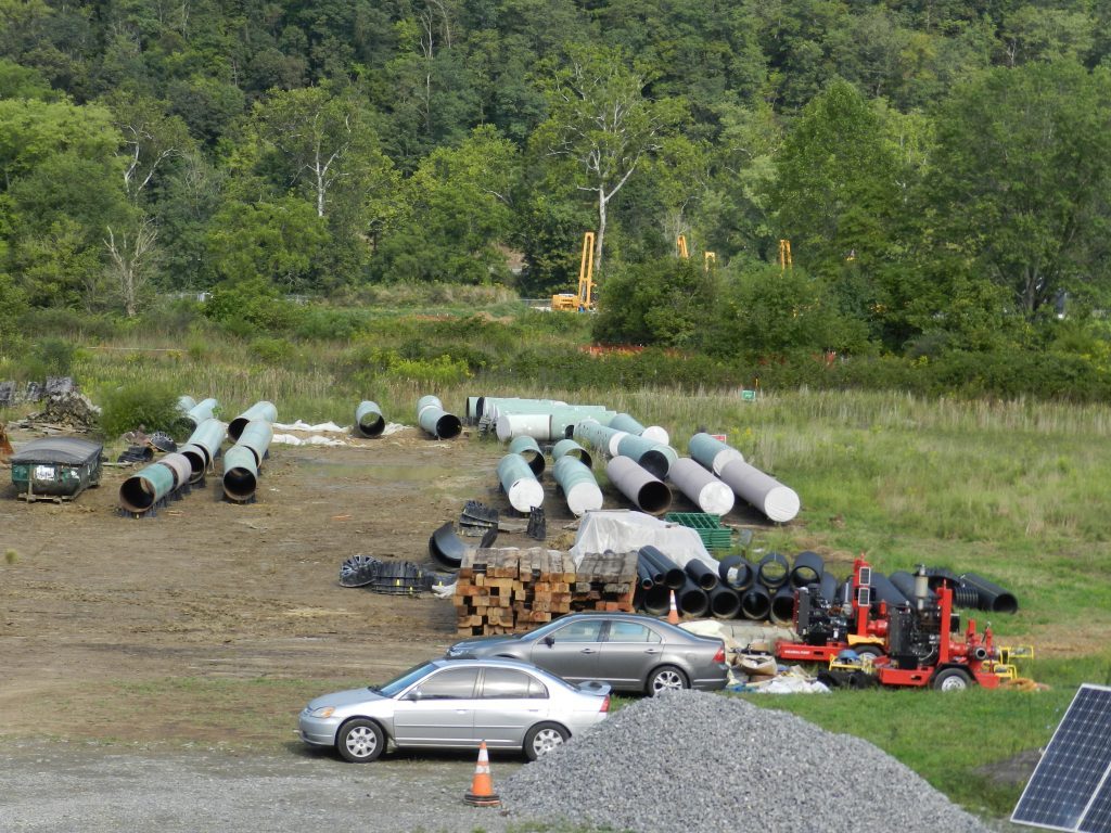Sections of large pipes lie exposed to to the elements in a cleared section of ground near a river. Large cranes loom over the river in the background. Cars, other equipment and a pile of gravel can be seen in the foreground.