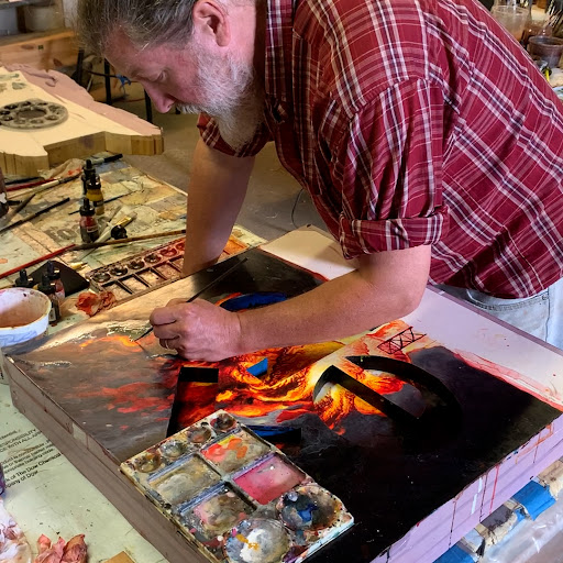A white-bearded man in a red and white plaid shirt and jeans leans over a canvas to work on a painting.