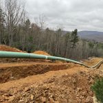 This lengthy section of green, pre-welded pipeline lays along side a deep, machine-dug trench that extends down a long slope through a cleared swath of forested land.