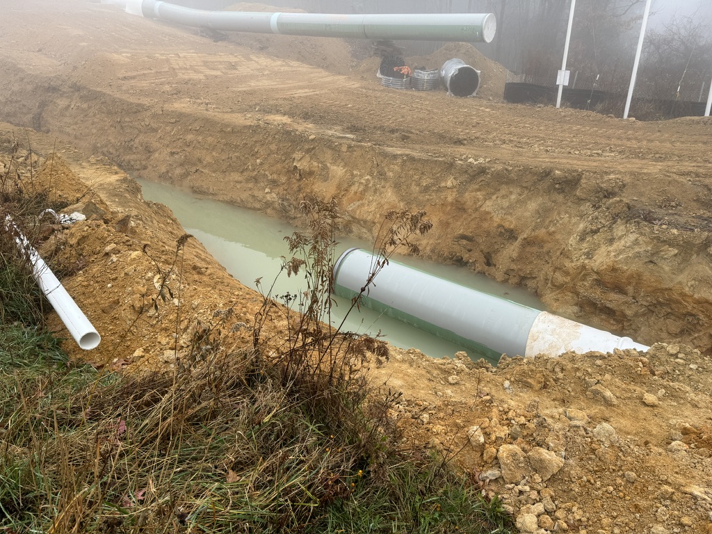 A large, partially buried, green pipe lays half submerged in water in a trench. Other pipe is visible off to the sides of the trench.