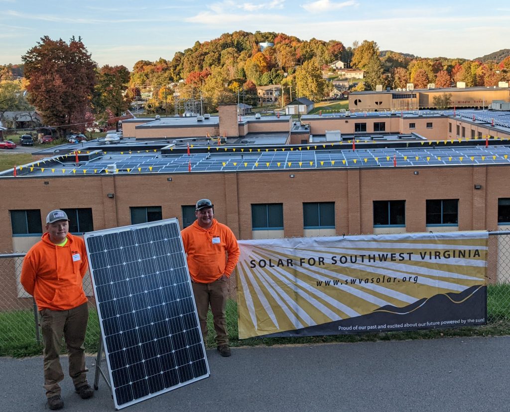two high school-aged boys inorange sweatshirts stan by a solar panel. A sign next to them says "Solar For Southwest Virginia" and behind them, solar panels cover the roof of a large brick building.