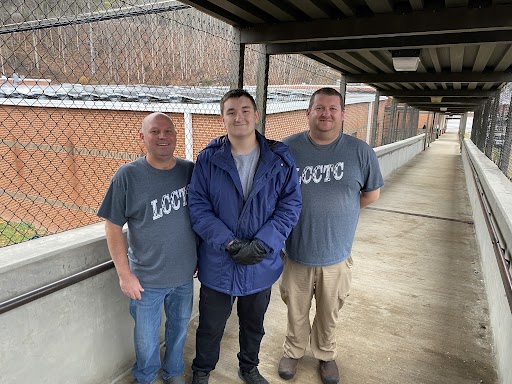 Three men stand on a covered walkway. The roof of the building behind them is covered by solar panels.
