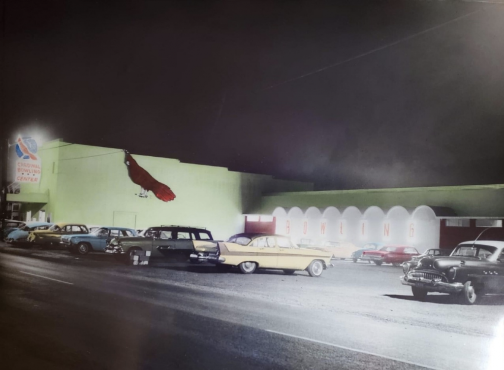 The parking lot outside a mint green painted building with a giant red cardinal on one wall is full of mid 20-th century cars. Signs designating it as Cardinal Lanes Bowling Center illuminate the nighttime scene.