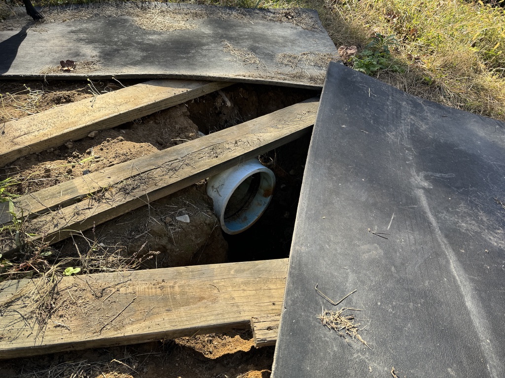 A drainage pipe sticks out into a drainage ditch. Planks and pieces of a dark material partially cover the ditch.