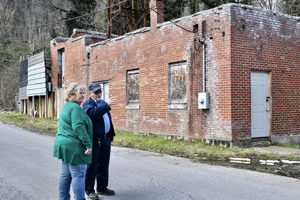 A woman in a green sweater and a man in a blue uniform stand beside a dilapidated brick building. They are looking toward the end of the road. The man is speaking.
