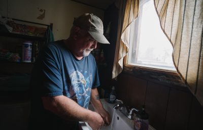 A white-bearded man in a blue t-shirt stands by a sink with water running from the faucet.