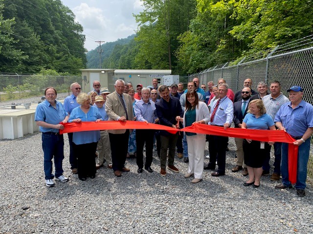 A large group of people stand behind a red ribbon on gravel in a fenced area in front of a small building. A man and woman in the front row of the group are cutting the ribbon with scissors.