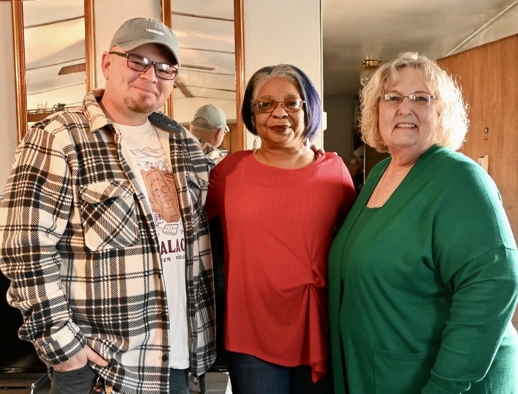 From the left, a man in a ball cap and plaid shirt stands next to a woman in a red shirt and a woman in a green shirt. They are standing inside a home and and smiling.