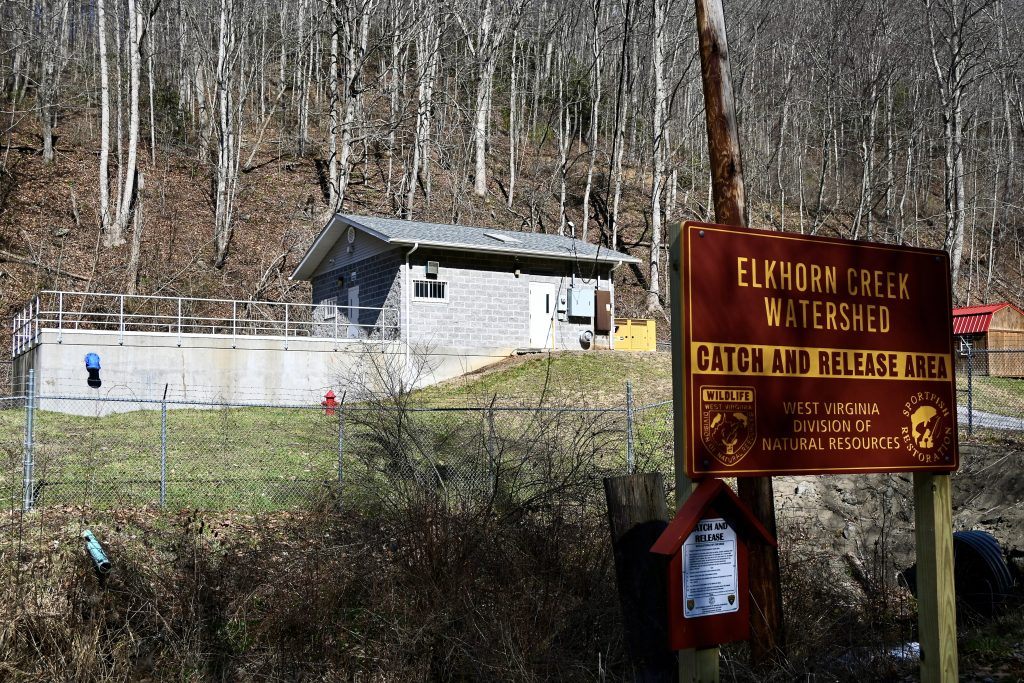 A cement block building sits behind a fence with barbed wire across the top. A sign in the foreground says "Elkhorn Creek Watershed, Catch and Release Area, West Virginia Division of Natural Resources."