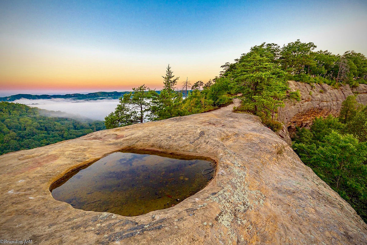 A depression in a rock ridge is filled with water. Clouds fill the valley below and sunrise colors the sky.