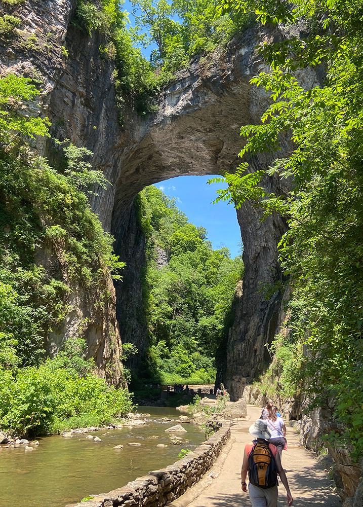 People walk along a path beside a river beneath a large stone arch.