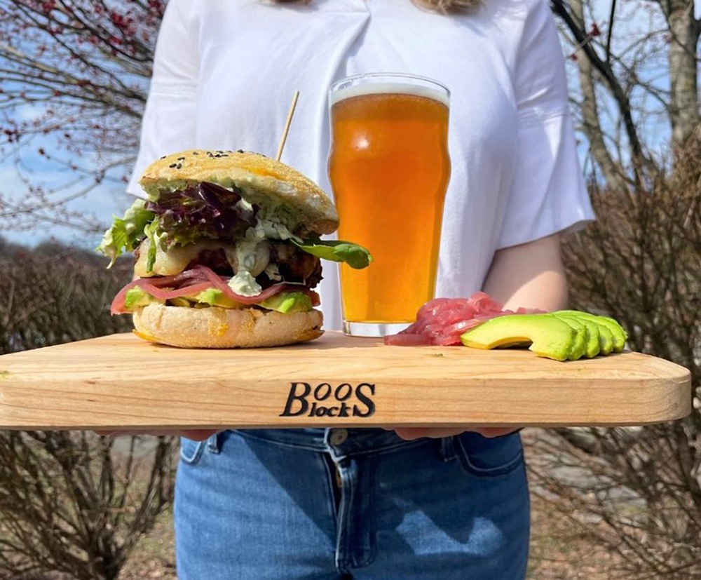 A person holds a large burger and beer on a wooden tray.
