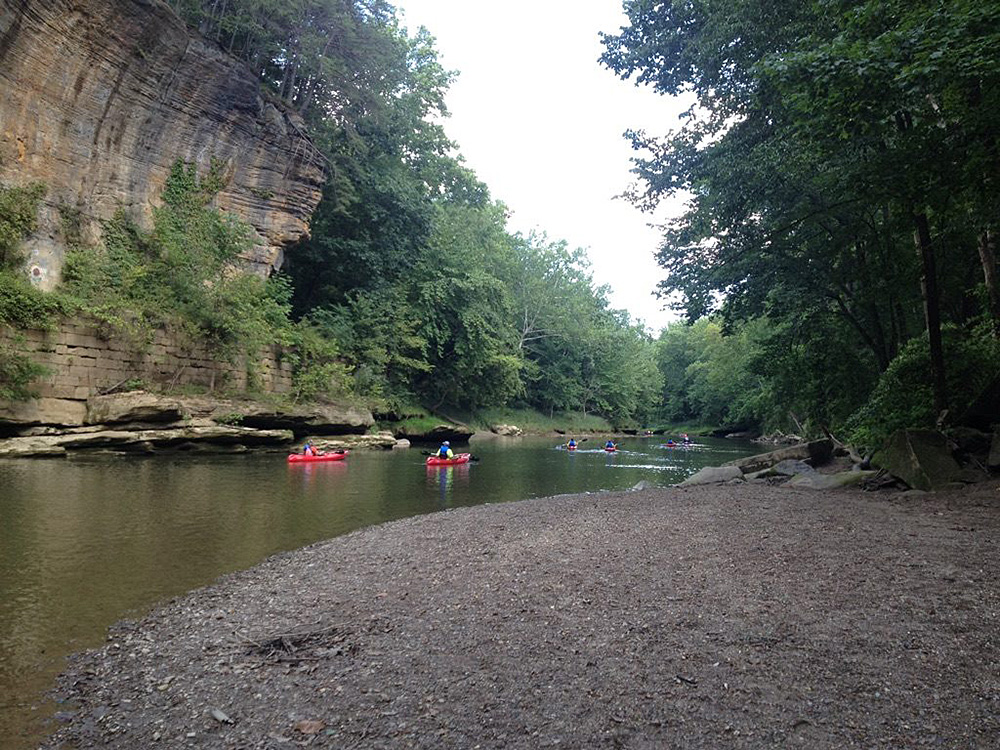 Two kayaks paddle on a river past a large rock wall