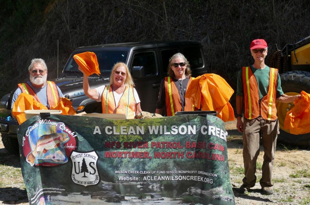 Four participants wearing orange vests, lifting trash bags and a "A Clean Wilson Creek" banner