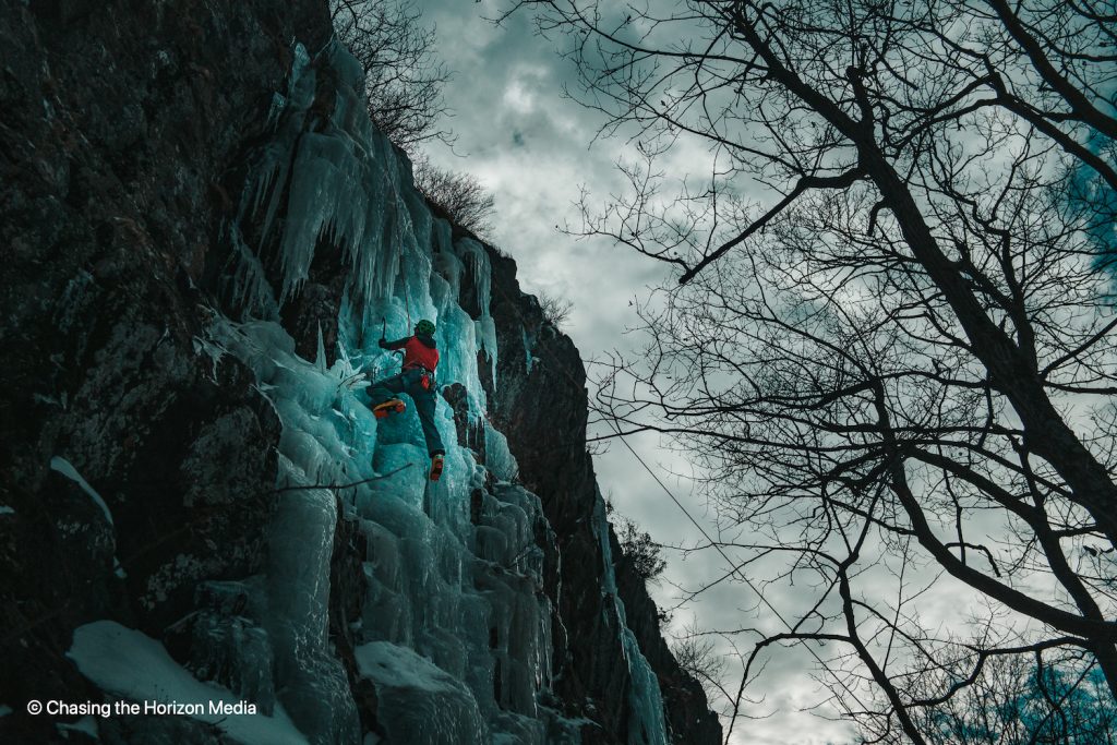 a man in a red jacket climbs a blue-tinged column of ice under a cloudy sky